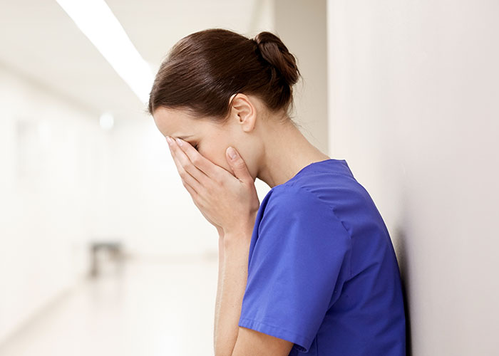 Female nurse in blue scrubs leaning against wall with hands covering face in an emotional moment in the ER hallway
