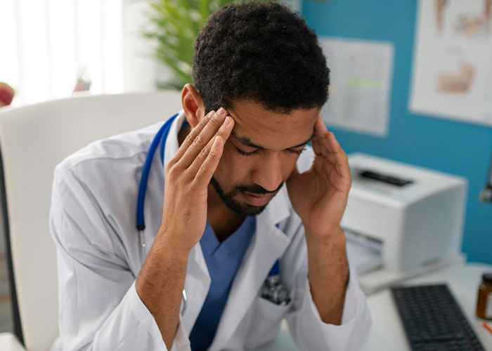 Doctor in a white coat with stethoscope holding head, appearing stressed while sitting in an ER office environment.