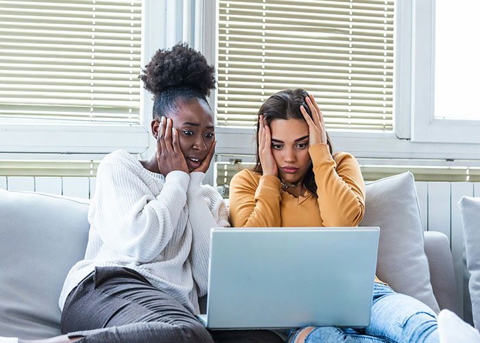 Two women sitting on a couch looking shocked and worried while reading disturbing threads about narrowly avoided kidnapping on a laptop.
