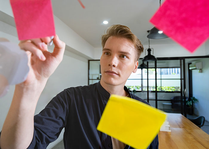 Young man arranging colorful sticky notes on glass wall, focused on planning a disturbing internet thread strategy.
