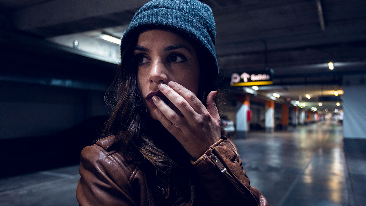Young woman in a beanie and leather jacket looking anxious alone in a dimly lit parking garage, evoking kidnapping fear.