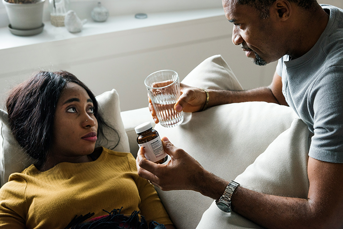 Man offering medication and glass of water to woman resting on couch during hospital recovery at home Man offering medication and glass of water to woman resting on couch during hospital recovery at home
