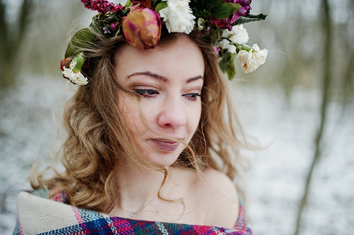 Woman wearing a flower crown and wrapped in a plaid shawl, looking sad outdoors, reflecting on paganism and family conflict.