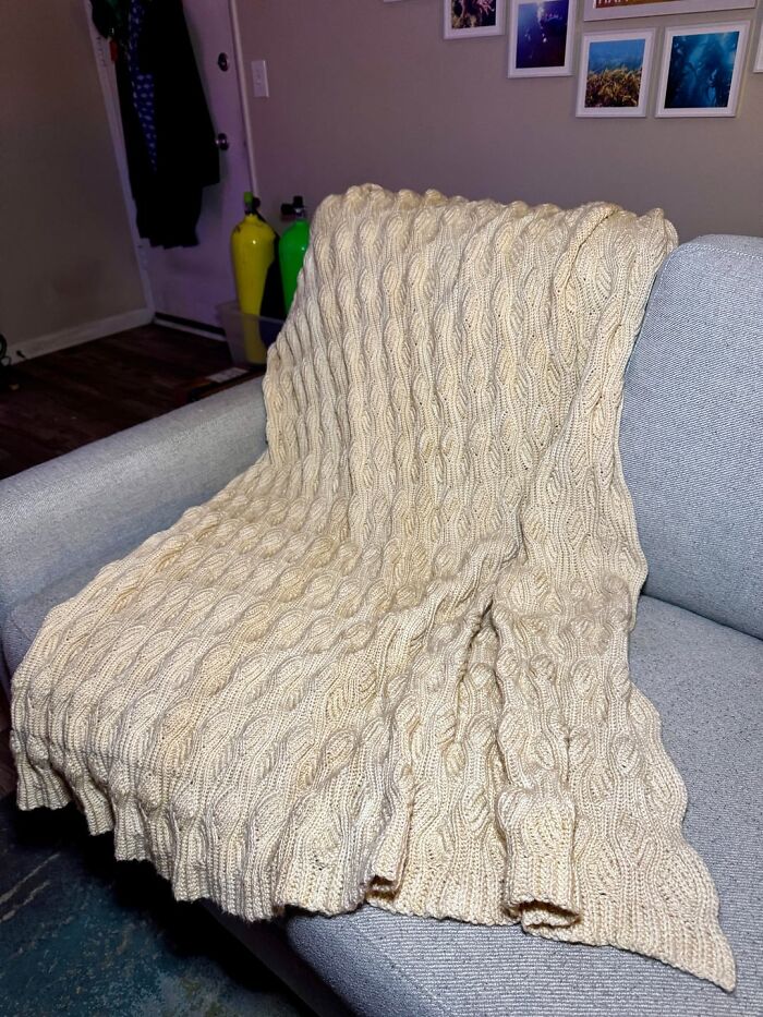 Cream-colored cable knit crochet blanket draped over a gray sofa in a cozy living room setting.