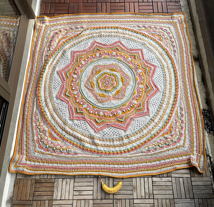 Crochet blanket with intricate floral and geometric patterns in white, pink, and yellow, laid out on wooden floor tiles.
