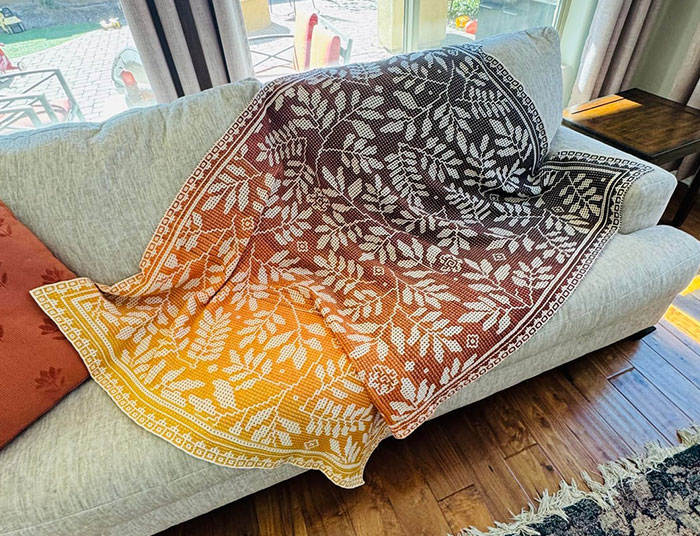 Intricately crocheted floral blanket with gradient colors draped over a modern gray sofa in a cozy living room.