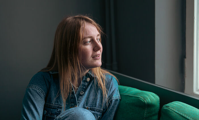 Young woman in a denim jacket sitting thoughtfully by a window, reflecting on feelings only adopted people understand.