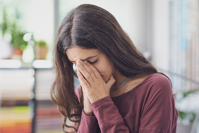Teen girl covering face with hands, appearing upset and distressed, sitting indoors in a bright room.