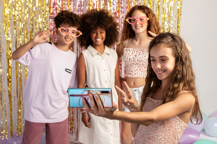 A 13-year-old girl taking a selfie with friends at a party, highlighting social drama and exclusion at school. A 13-year-old girl taking a selfie with friends at a party, highlighting social drama and exclusion at school.