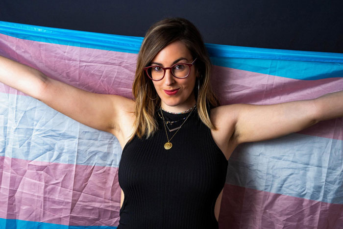 Young woman with glasses posing confidently with a trans pride flag, symbolizing daughter money and surgeries topics. Young woman with glasses posing confidently with a trans pride flag, symbolizing daughter money and surgeries topics.