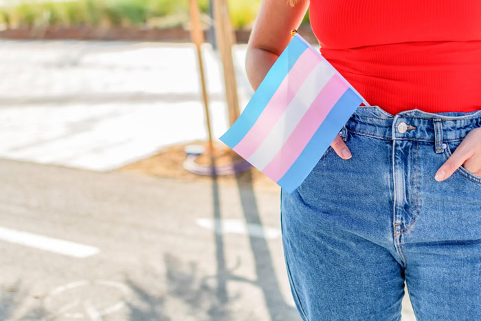 Person wearing a red top and jeans holding a transgender pride flag representing daughter money surgeries and support. Person wearing a red top and jeans holding a transgender pride flag representing daughter money surgeries and support.