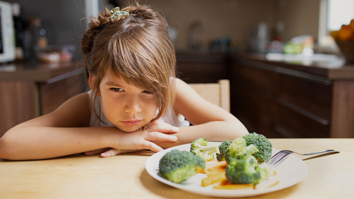 Young girl in kitchen looking upset at plate of broccoli and vegetables, reflecting foodie woman and kid conflict.