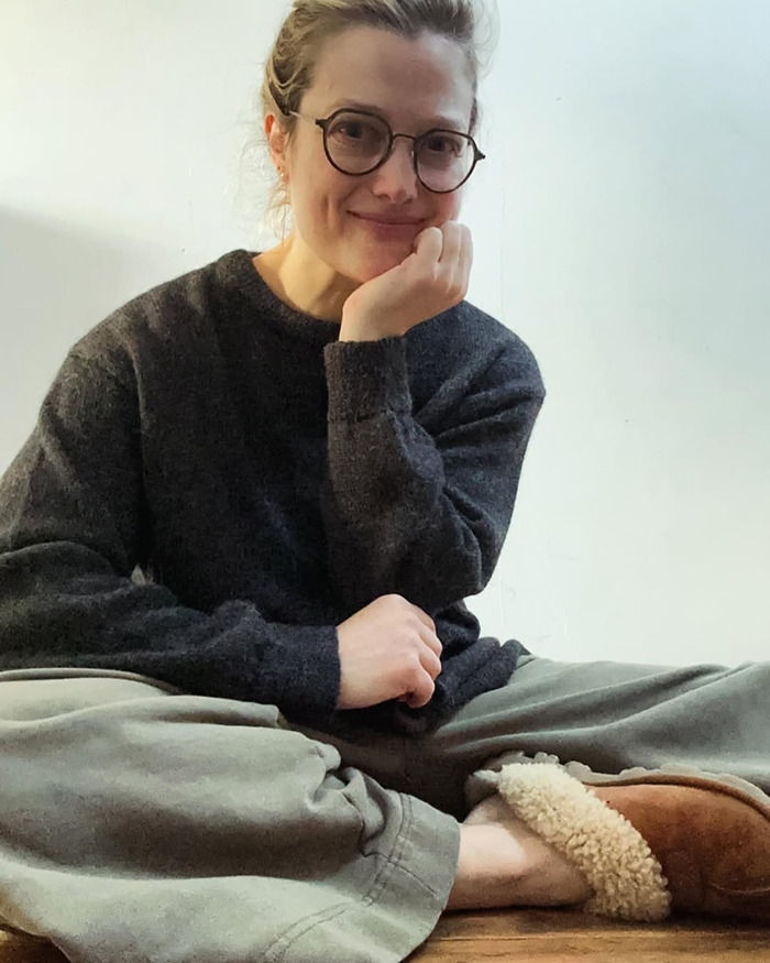 Alison Sudol sitting casually indoors wearing glasses, a dark sweater, and cozy slippers, smiling gently at the camera Alison Sudol sitting casually indoors wearing glasses, a dark sweater, and cozy slippers, smiling gently at the camera