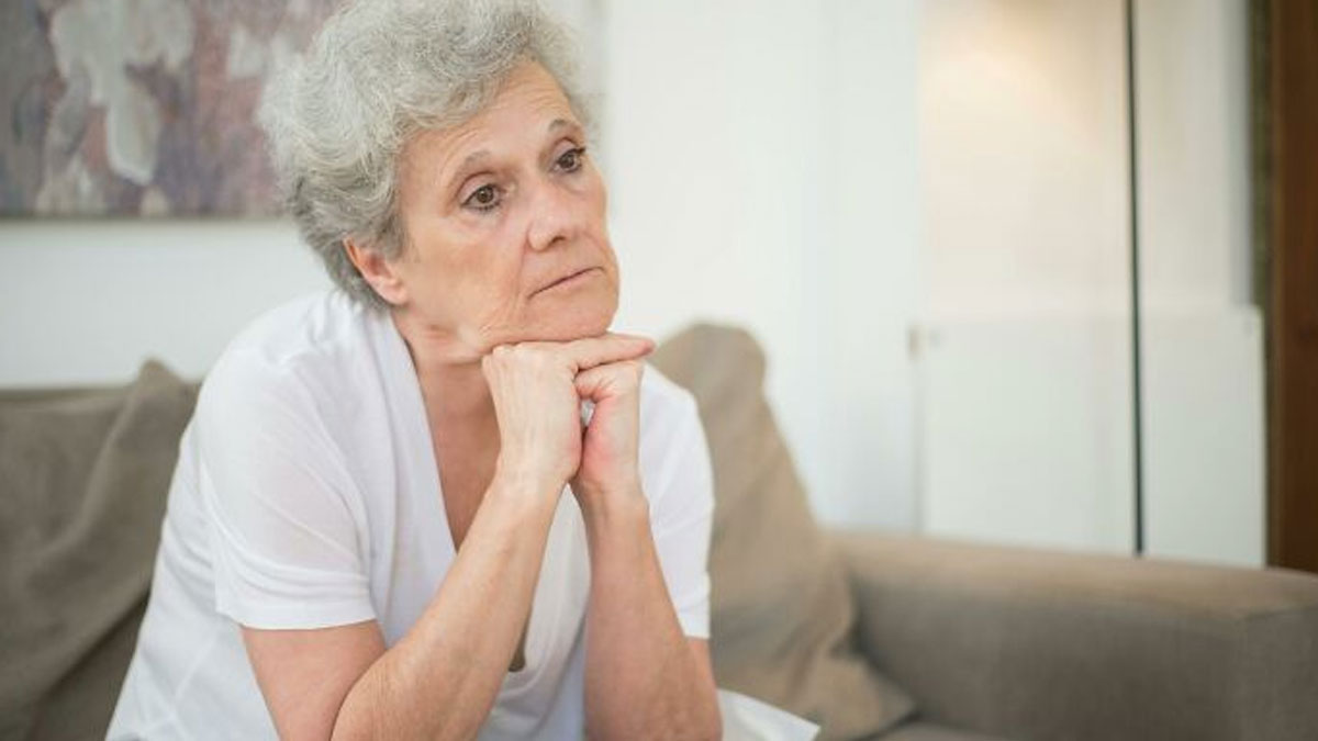Elderly woman sitting on a couch, looking thoughtful and upset about not seeing her grandkids.