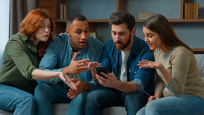 Four young adults sitting on a couch, looking shocked while viewing a phone, illustrating family health fund dispute. Four young adults sitting on a couch, looking shocked while viewing a phone, illustrating family health fund dispute.