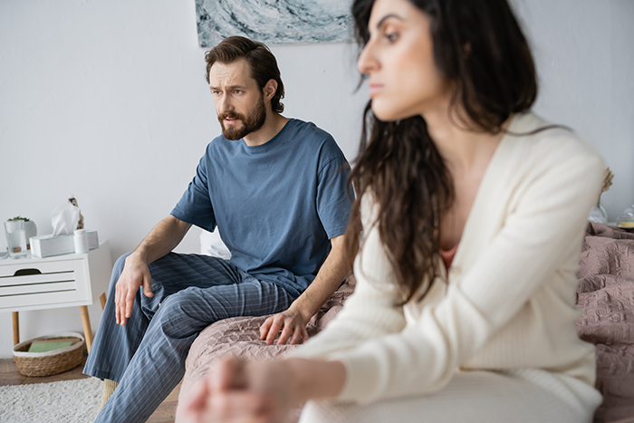 A couple sits apart on a bed, showing tension after a confrontation about Thanksgiving food hogging by MIL.