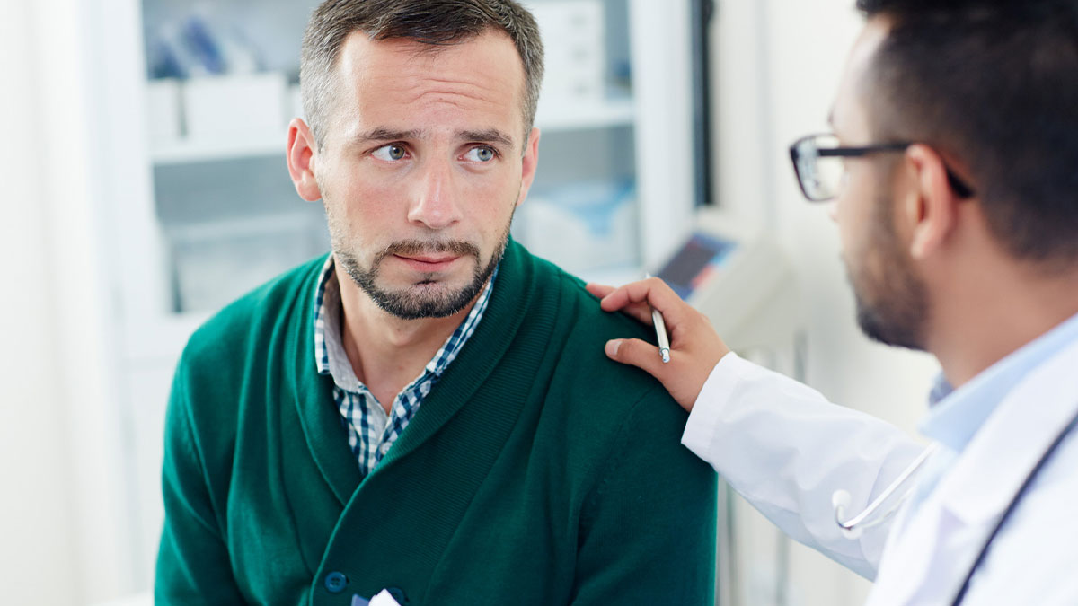 Male patient looking concerned while a doctor wearing glasses offers support with a hand on his shoulder in a clinic.