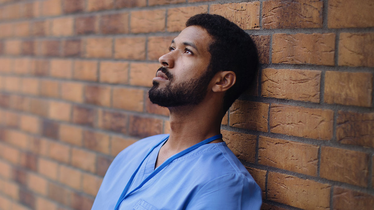 Surgeon in blue scrubs leaning against a brick wall, appearing thoughtful and reflecting on past mistakes.