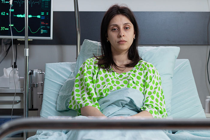 Young woman in a hospital bed wearing a green polka dot gown, connected to medical monitors after doctors messed up bad.