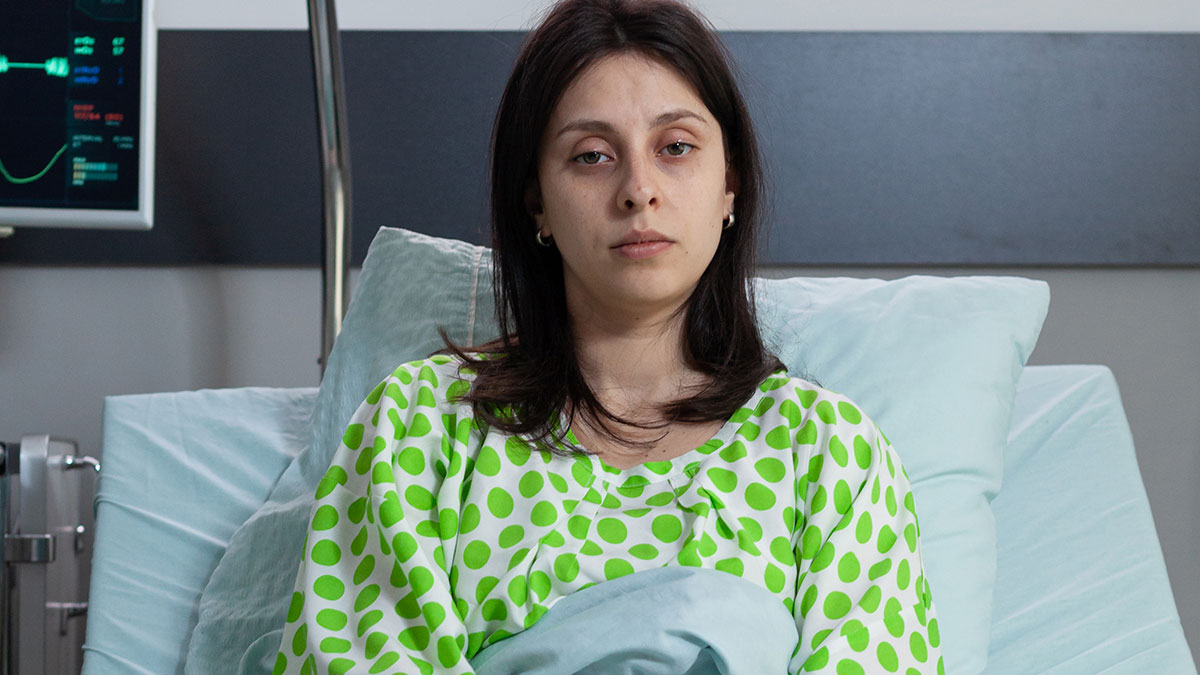 Woman in hospital bed wearing green polka dot gown, looking tired and unwell after doctors messed up medical care.