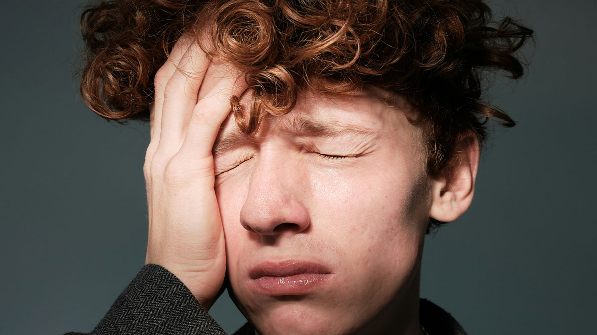 Young man with curly hair facepalming, expressing disbelief, illustrating Americans confidently posting dumbest things.