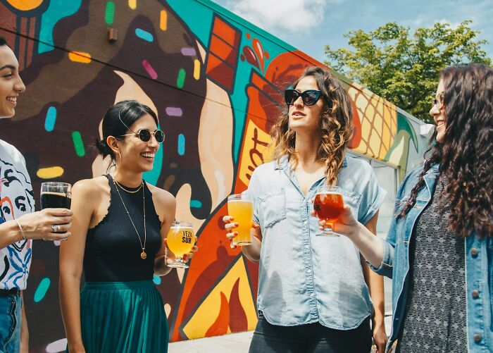 Four women laughing and holding drinks outdoors by a colorful mural, representing ways people found out unfaithful partners.