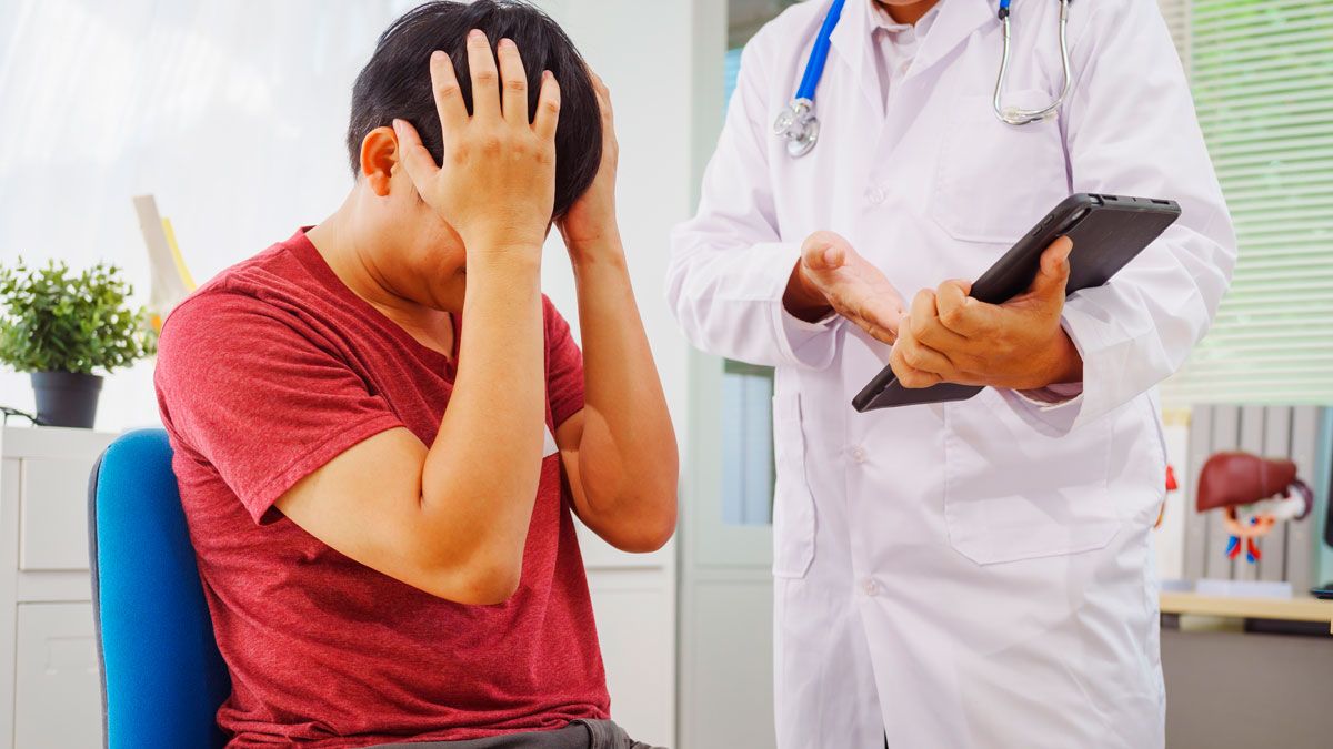 Man holding his head in embarrassment during a doctor visit while healthcare professional reviews notes on a tablet.