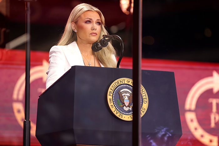 Erika Kirk speaking emotionally behind a podium with the presidential seal during a public address.