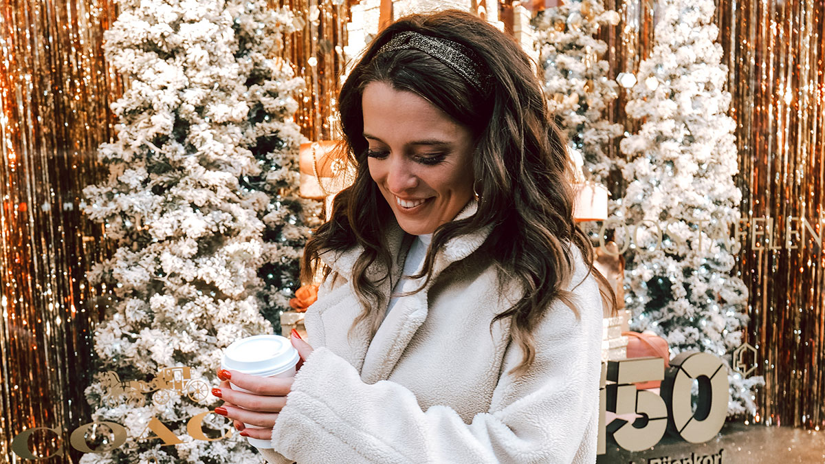 Woman in a cozy coat holding a coffee cup in front of decorated snowy Christmas trees at a European Christmas market.