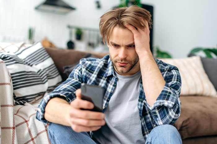 Man sitting on couch looking stressed while holding phone, reflecting on refusing to visit terminally ill ex-GF who cheated. Man sitting on couch looking stressed while holding phone, reflecting on refusing to visit terminally ill ex-GF who cheated.