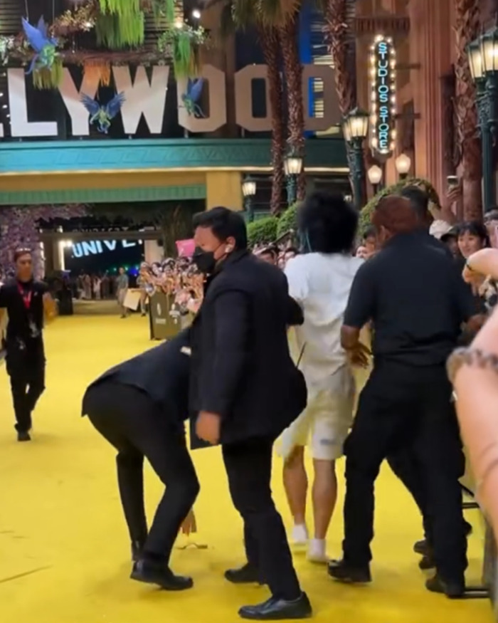 Security personnel restraining a fan who attacked Ariana Grande at a premiere as crowd looks on and records the event. Security personnel restraining a fan who attacked Ariana Grande at a premiere as crowd looks on and records the event.