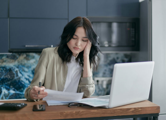 Young woman looking stressed reviewing documents at table with laptop, relating to guy charging daughter $15K for raising her. Young woman looking stressed reviewing documents at table with laptop, relating to guy charging daughter $15K for raising her.