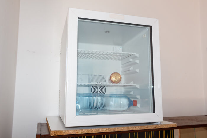 Small white fridge with broken shelves and a single water bottle seen through the glass door inside a sparse room. Small white fridge with broken shelves and a single water bottle seen through the glass door inside a sparse room.