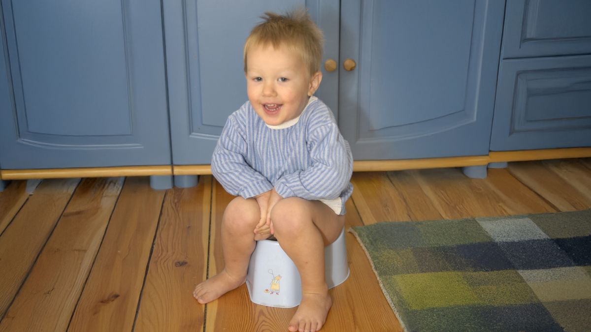 Toddler sitting on a potty chair smiling in a kitchen, capturing the fun side of p**p jokes for kids.