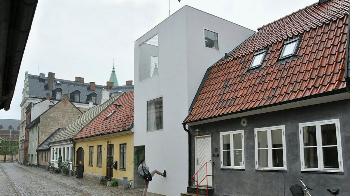 A narrow, unesthetic building with modern architecture contrasts with older traditional houses along a cobblestone street.