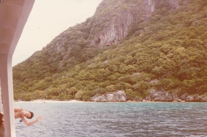 Man photobombing from boat window with lush green mountains and clear blue water in the background.