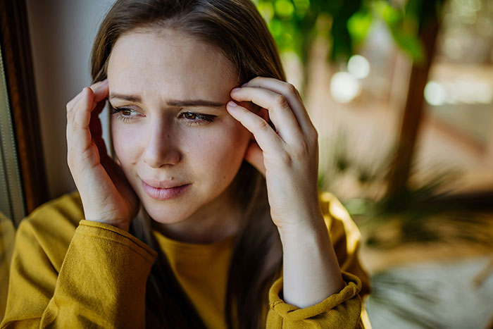 Woman in yellow sweater looking troubled by window, reflecting on attempts to drive a wedge between brother and his wife.