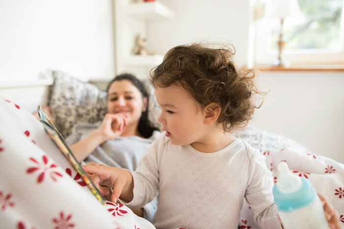 Young child reading a book while mother relaxes on bed, illustrating family dynamics in a couple's move for DIL job.