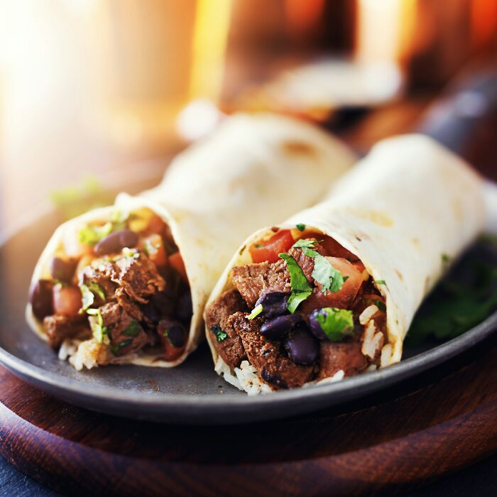 Close-up of two burritos filled with beef, black beans, rice, and fresh vegetables on a dark plate, food etymology concept.