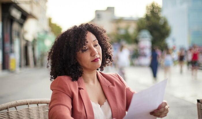 Woman with curly hair in a salmon blazer looking thoughtfully at a paper outdoors, illustrating uniquely awkward wedding moments.