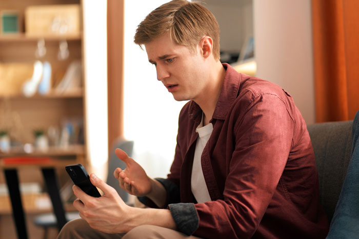 Young man looking irked while holding phone, sitting indoors, reflecting on friend who likes him after crying incident. Young man looking irked while holding phone, sitting indoors, reflecting on friend who likes him after crying incident.