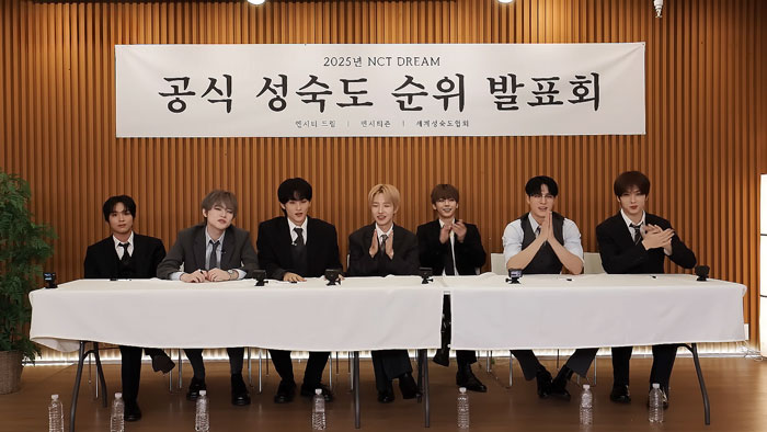 K-Pop band NCT DREAM dressed formally, seated at a panel table with microphones and water bottles on the floor. K-Pop band NCT DREAM dressed formally, seated at a panel table with microphones and water bottles on the floor.