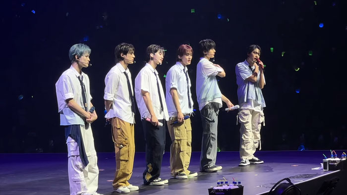 K-pop band performing on stage with six members in casual outfits during a live concert event.