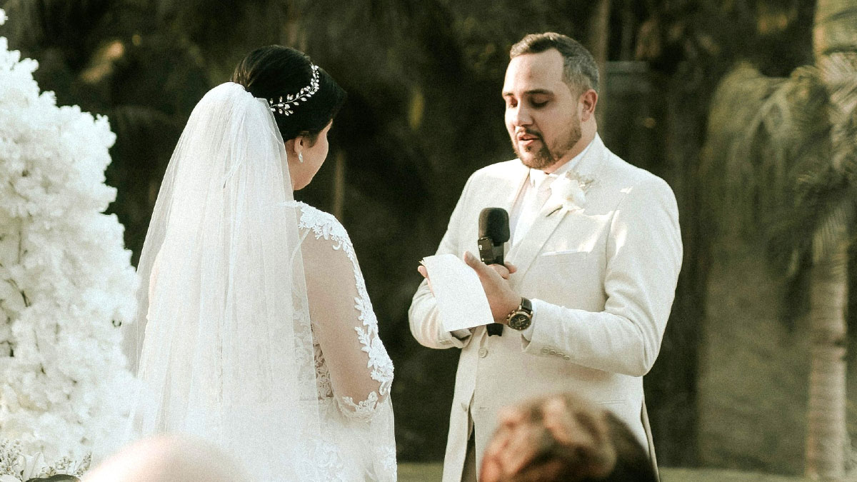 Groom in white suit reading wedding vows outdoors to bride in lace dress with veil during ceremony with microphone.