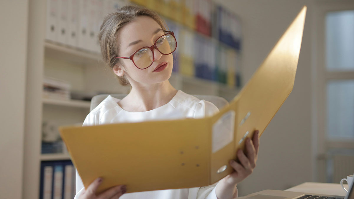 Young woman wearing glasses reviewing a large yellow folder at her desk, related to mocking fiancu00e9eu2019s job story.