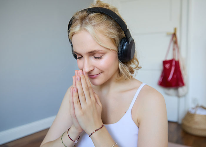 Young woman practicing unusual wellness habits, wearing headphones and meditating with eyes closed indoors.
