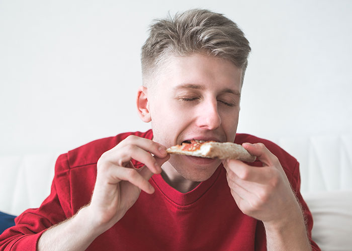 Young man enjoying pizza with eyes closed, illustrating unusual wellness habits experts swear by.