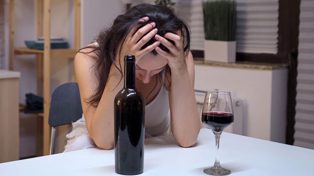 Woman with head in hands sitting at table with wine bottle and glass, showing distress and failed life signs.