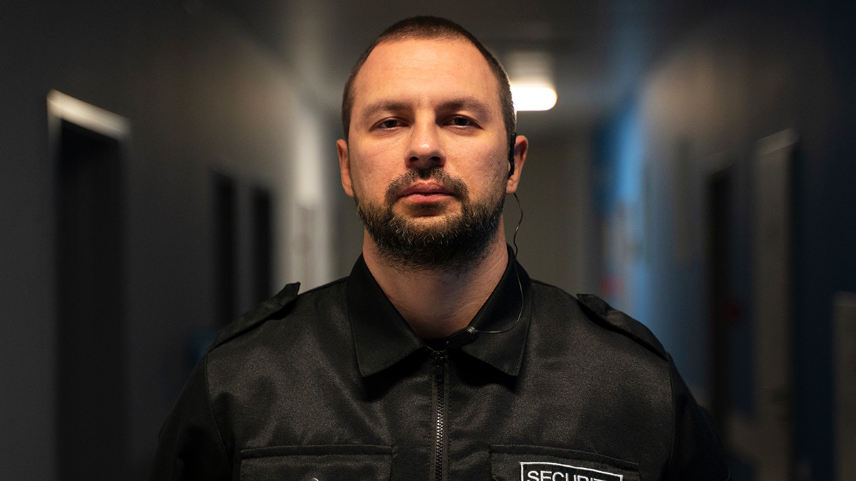 Man in security uniform with earpiece standing in a dimly lit hallway, reflecting corporate comedy gold theme.