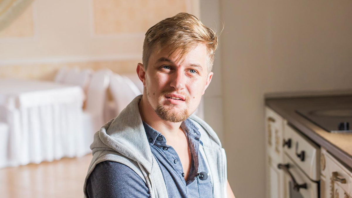 Young man in a casual shirt and hoodie sitting indoors, expressing emotional struggle while caring for disabled brother.